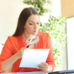 Frau liest Dokument mit Lupe am Schreibtisch.