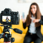 Frau filmt sich selbst mit Kamera, während sie auf Smartphone zeigt, im Wohnzimmer.