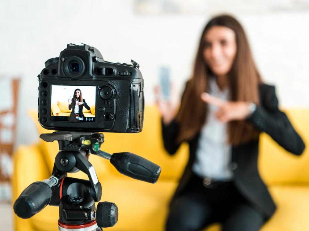 Frau filmt sich selbst mit Kamera, während sie auf Smartphone zeigt, im Wohnzimmer.